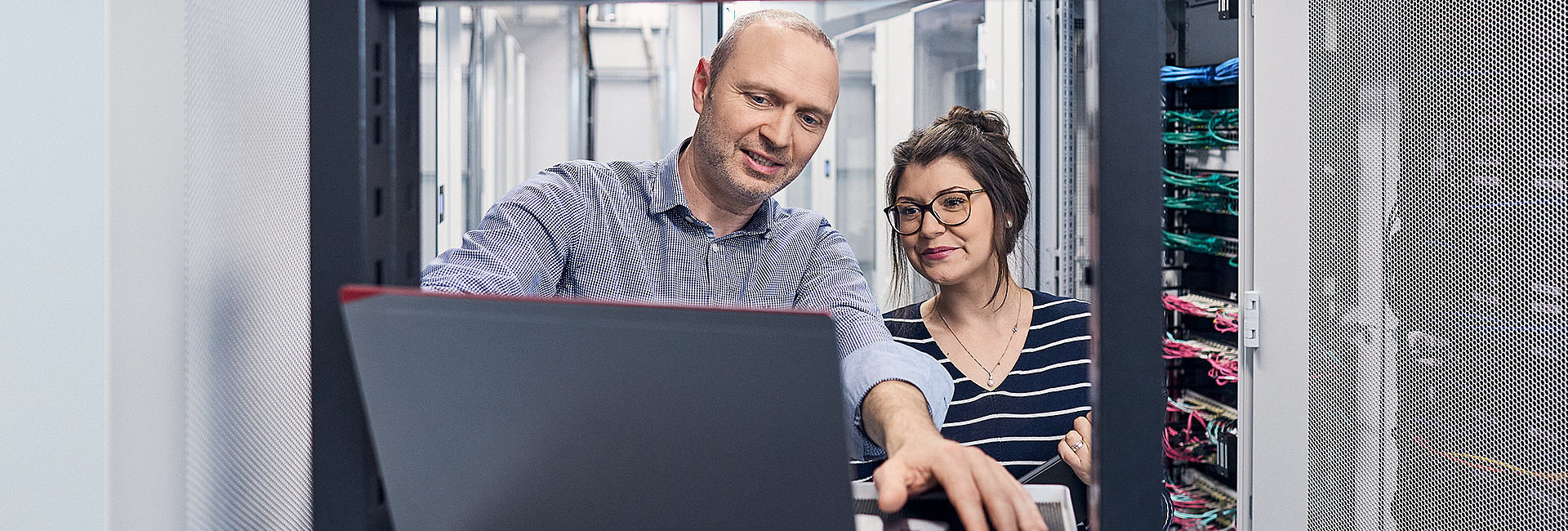 Frau und Mann schauen in einem Server Raum zusammen auf einen Laptop