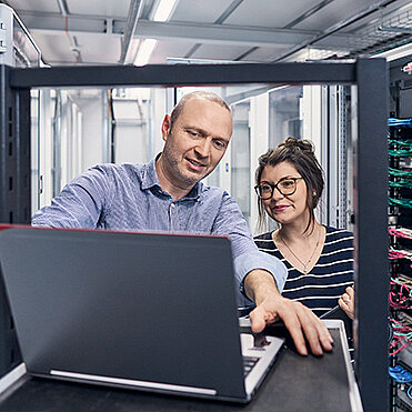 Frau und Mann schauen in einem Server Raum zusammen auf einen Laptop