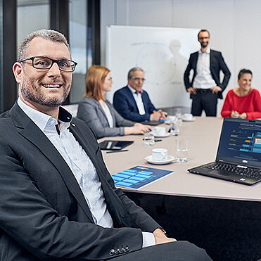 Eine Besprechung unter Mitarbeitern, im Vordergrund ein Mann, der in die Kamera schaut