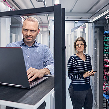 Mann und Frau schauen in einem Server Raum auf einen Laptop